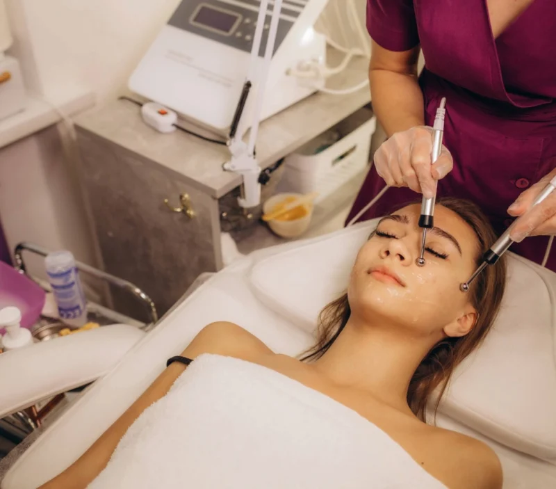 A technician is performing a CACI facial on a young woman lying on a spa bed. The technician uses two handheld probes to apply a procedure to the woman's face, specifically near her eyes.