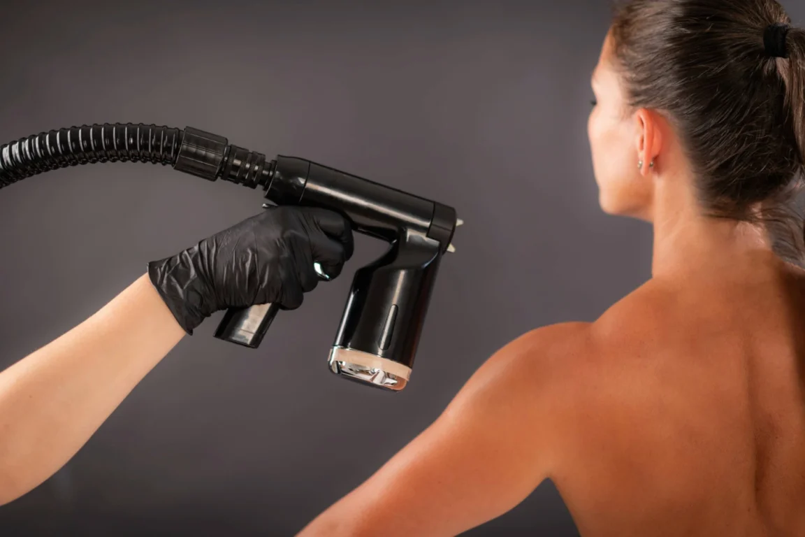 Spray tan specialist pointing fake tan spray gun at client’s back.